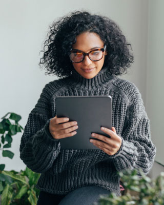 A person with curly hair and glasses is seated indoors, wearing a cozy gray sweater, and reading a tablet. There are some green plants in the background, adding a touch of nature to the setting.