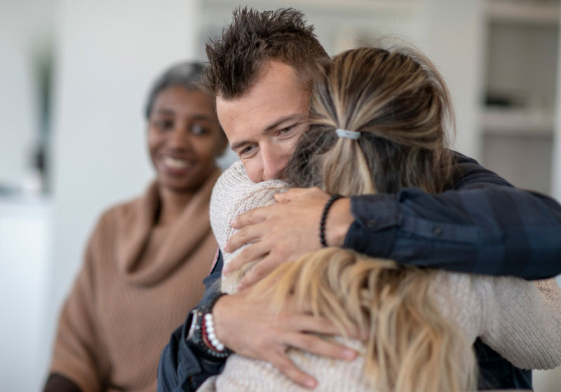 A man and a woman share a warm hug, displaying a sense of support and comfort. A smiling woman sits in the background, observing the interaction. The scene conveys an atmosphere of compassion and connection.