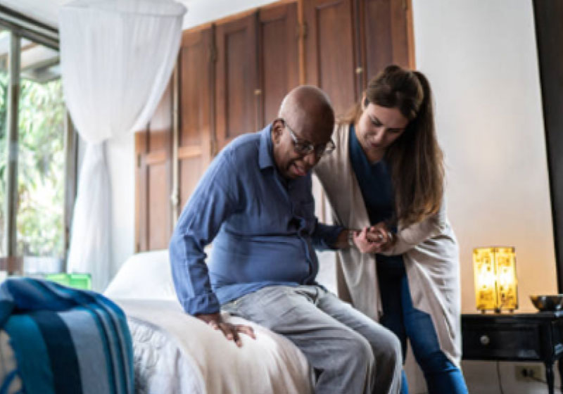 A young woman assists an elderly man as he sits on the edge of a bed in a well-lit room with wooden wardrobes in the background. The woman is helping the man stand up, holding his hand and supporting his back.