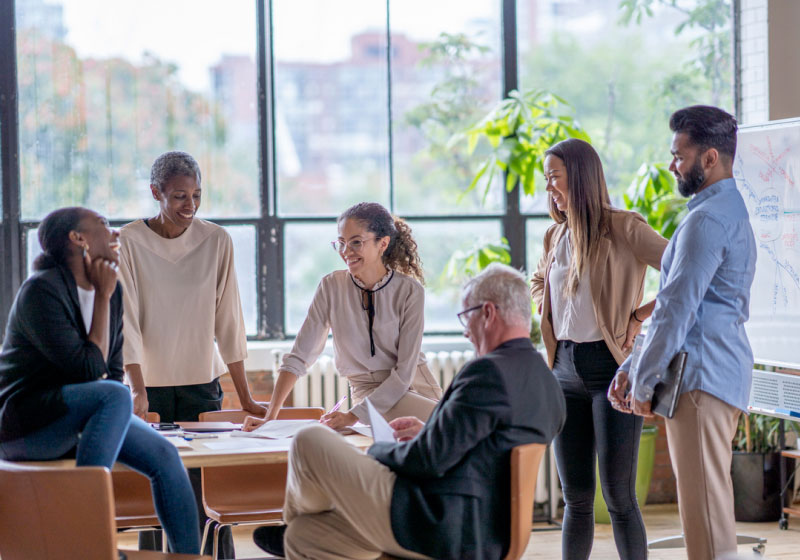 A group of five people engaged in a lively discussion in a bright office space with large windows. They are gathered around a table with documents, and a whiteboard with diagrams is visible in the background. One person sits while the others stand or lean casually.