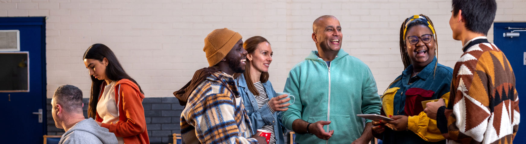 A group of people of various ages and ethnicities are gathered indoors, engaging in conversation and smiling. Some hold cups or plates. They are dressed in casual, colorful clothing. A light brick wall and blue doors are visible in the background.