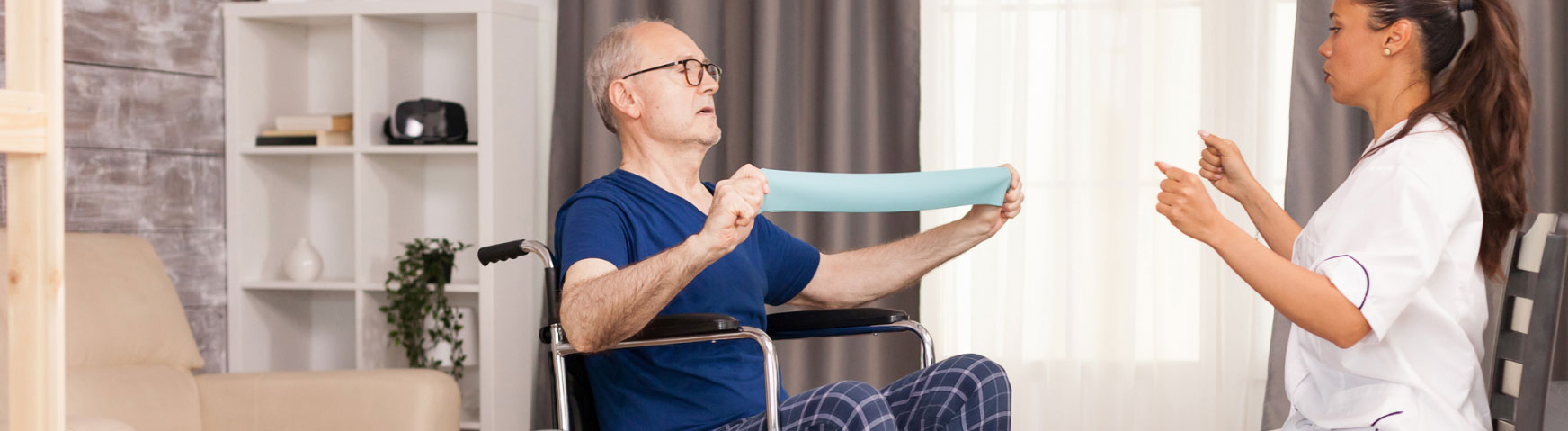 A senior man in a wheelchair exercises using a resistance band, assisted by a female caregiver in a white uniform. They are in a well-lit living room with a shelf and curtains in the background.