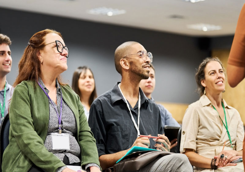 A diverse group of people sitting in a brightly lit conference room attentively listening to a speaker. The audience members are smiling and appear engaged, with some taking notes. They wear name tags and are dressed in casual to business casual attire.