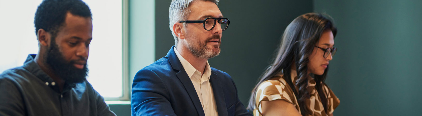 Three people are seated at a table, appearing focused and engaged. The person on the left has a beard and wears a dark shirt, the person in the middle wears glasses and a blue suit, and the person on the right has long hair and wears a patterned top.