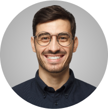 A man with short dark hair, a trimmed beard, and round glasses smiles at the camera. He is wearing a dark-colored button-up shirt. The background is a gradient of light gray. The image is cropped in a circular frame.