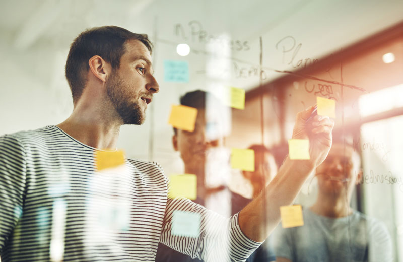 A man in a striped shirt is placing a sticky note on a glass wall filled with various other brightly colored sticky notes during a collaborative meeting. Two colleagues stand in the background, partially visible and engaged in the discussion.