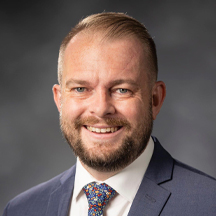 Kevin Shafer, a man with short blond hair and a beard, wearing a dark suit jacket, white shirt, and colorful patterned tie smiles in front of a blurred gray background.