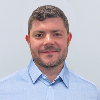 A man with short, curly dark hair and a beard smiles at the camera. He is wearing a light blue collared shirt against a plain, light-colored background.